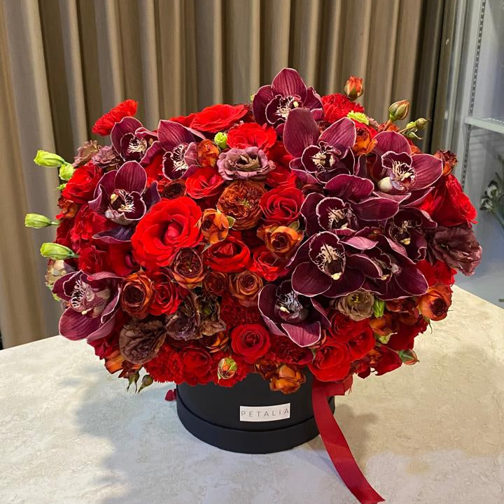 A large, dense round floral arrangement featuring red and burgundy roses, red orchids, and small orange-brown flowers, presented in a black round box with a red ribbon on a light marble tabletop.